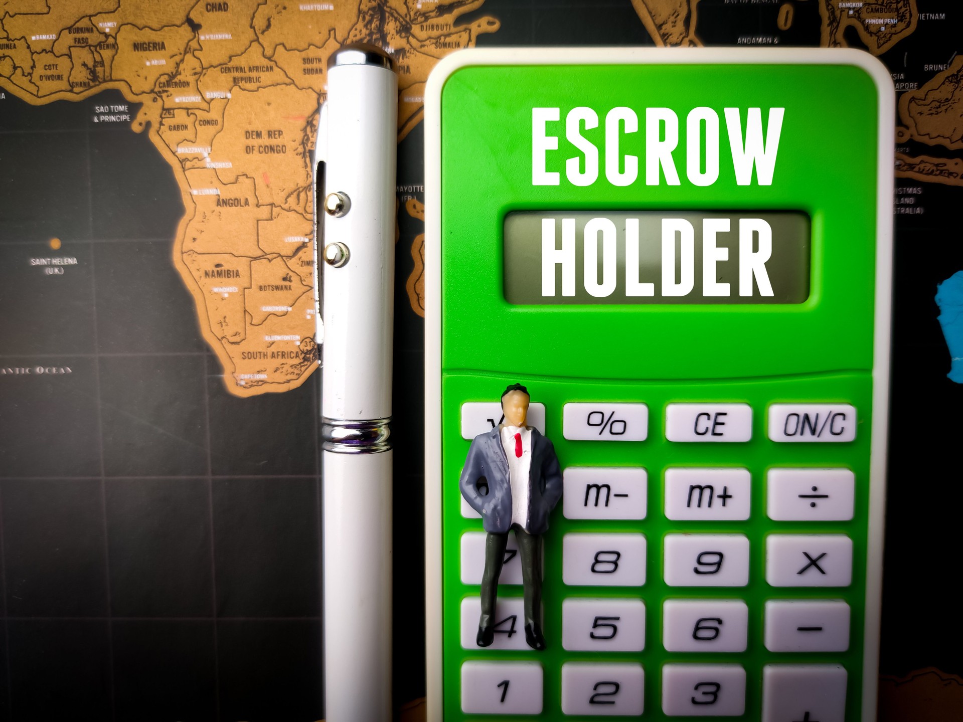 Calculator,miniature people and pen with text ESCROW HOLDER on a world map background